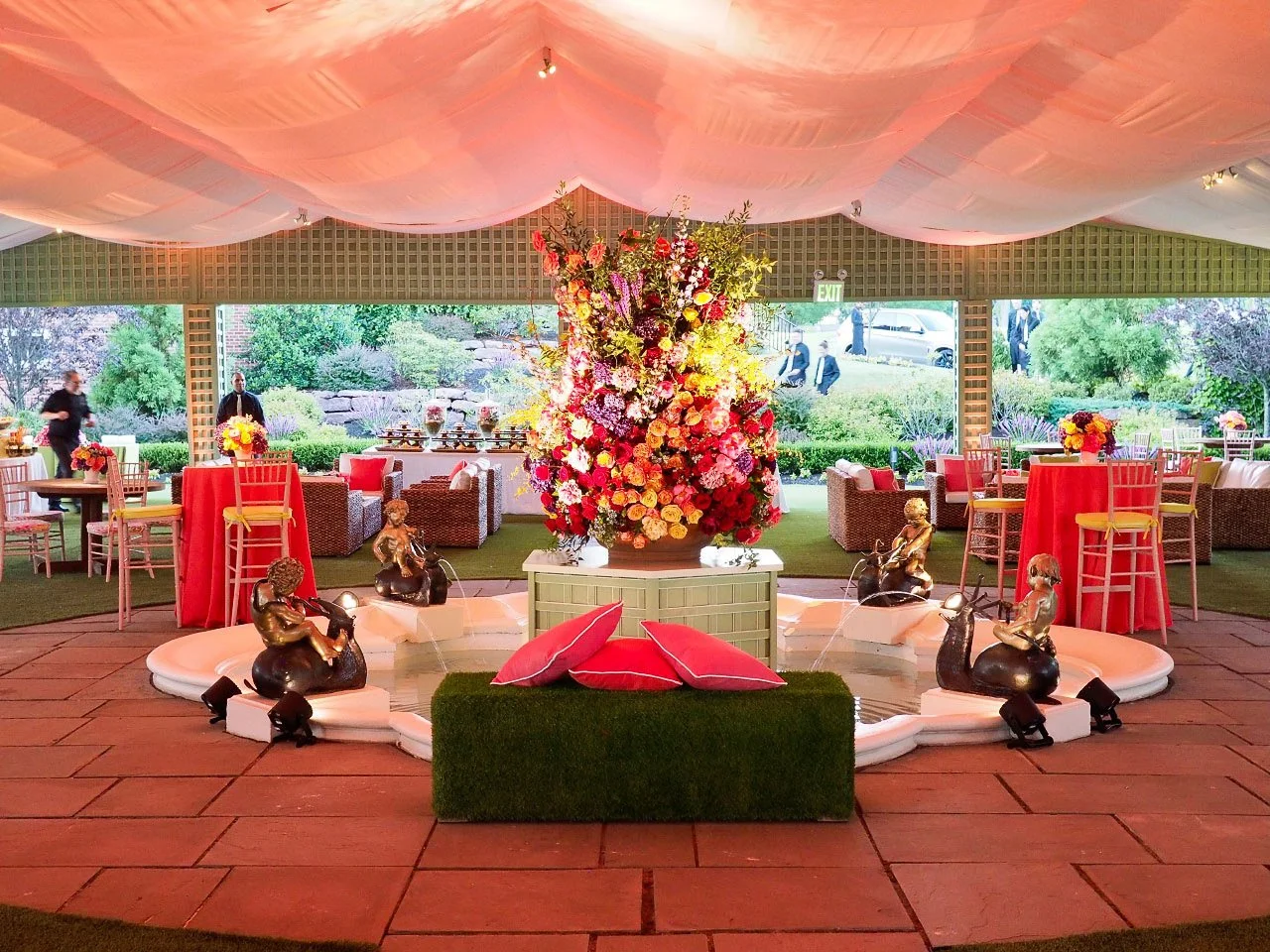 Elegant event space with a large colorful floral centerpiece on a green pedestal, surrounded by pink and red cushions, gold cherub sculptures, and seating areas with tables and floral arrangements. The ceiling is draped with pink fabric, and large wi