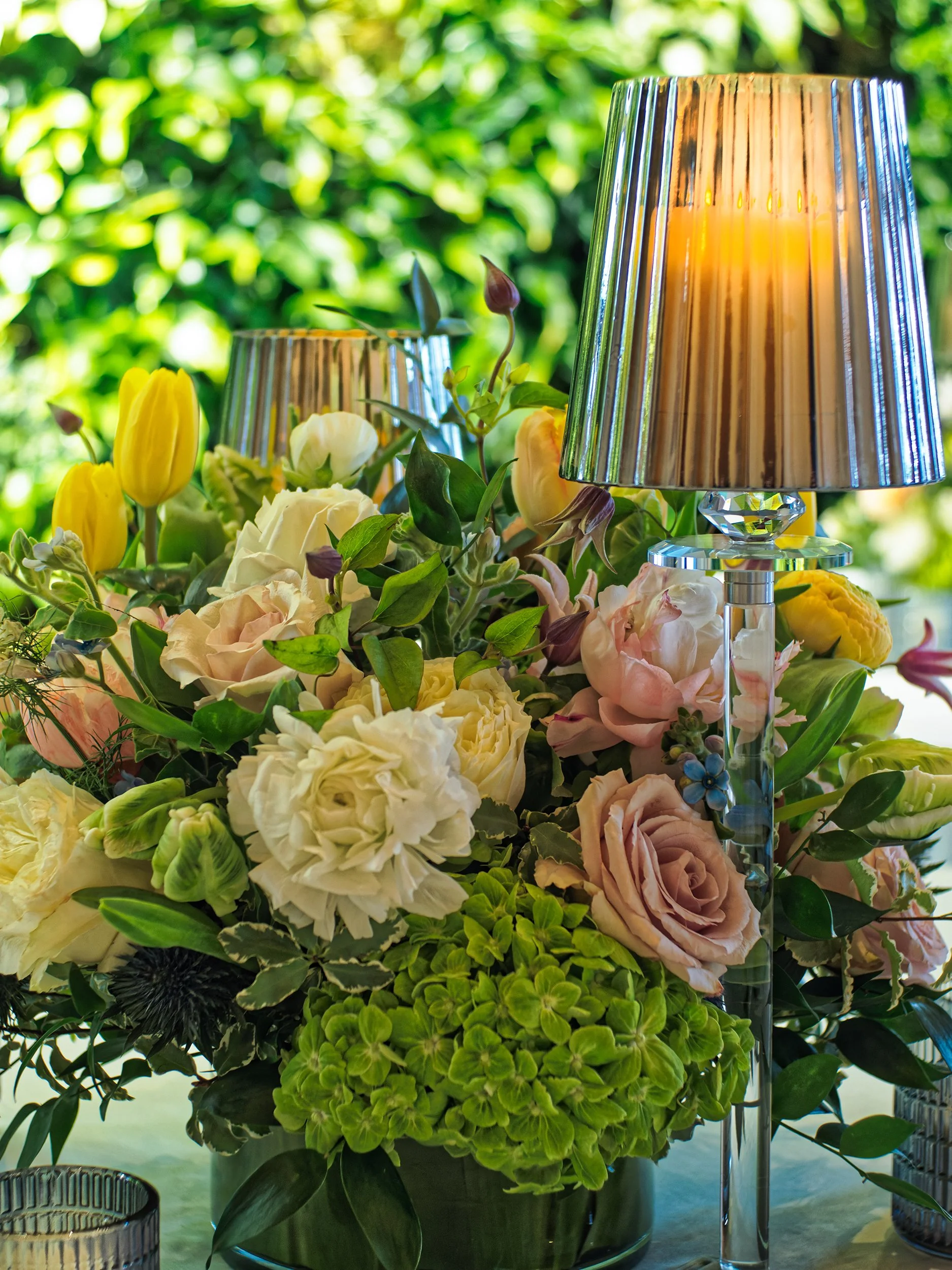 A vibrant flower arrangement with pink, white, yellow, and purple flowers, surrounded by green foliage, with two tall, metallic lamps with pleated shades in the background.