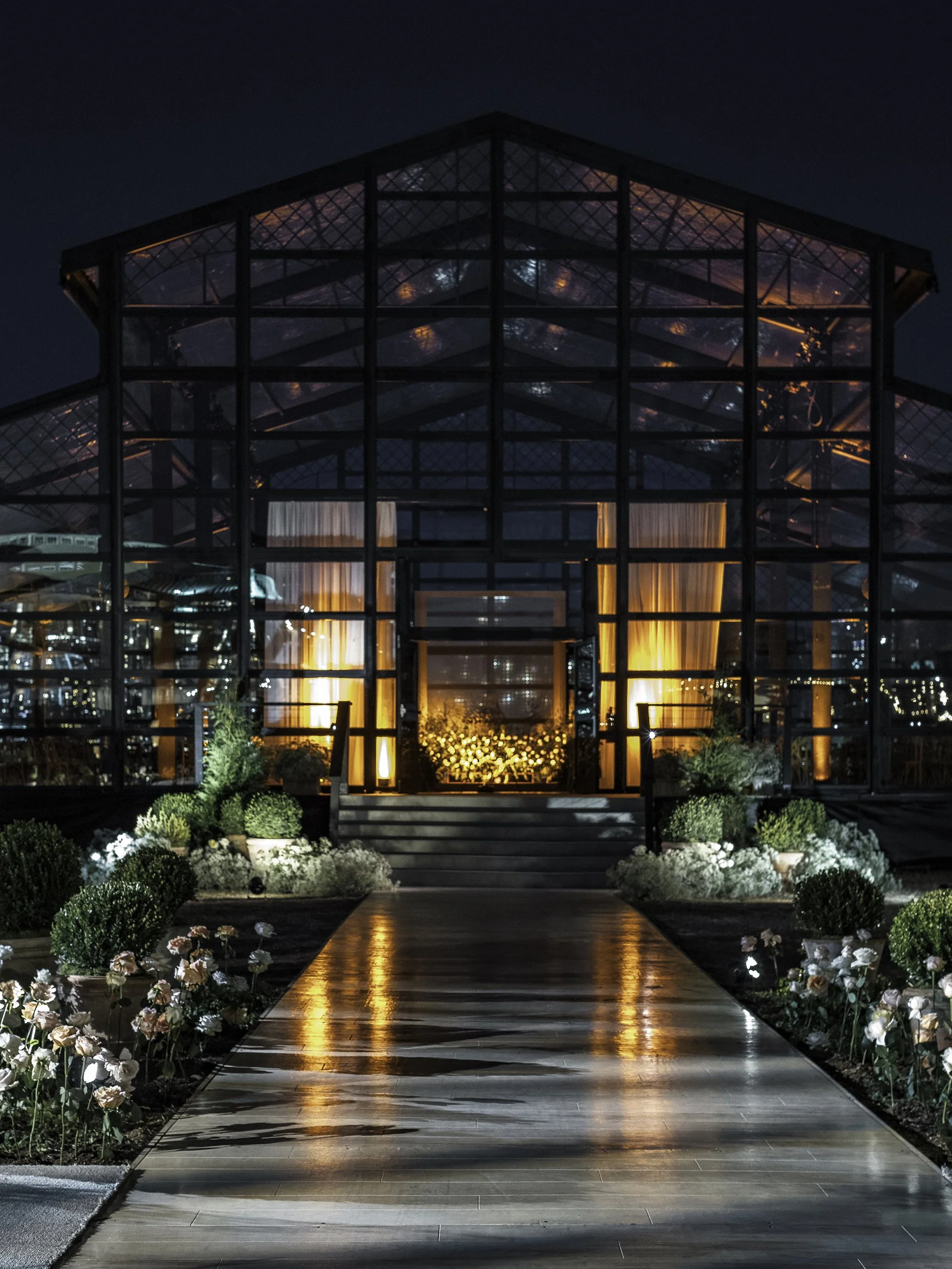 Night view of a glass greenhouse with warm interior lighting, a pathway with reflected lights, and floral decorations outside.