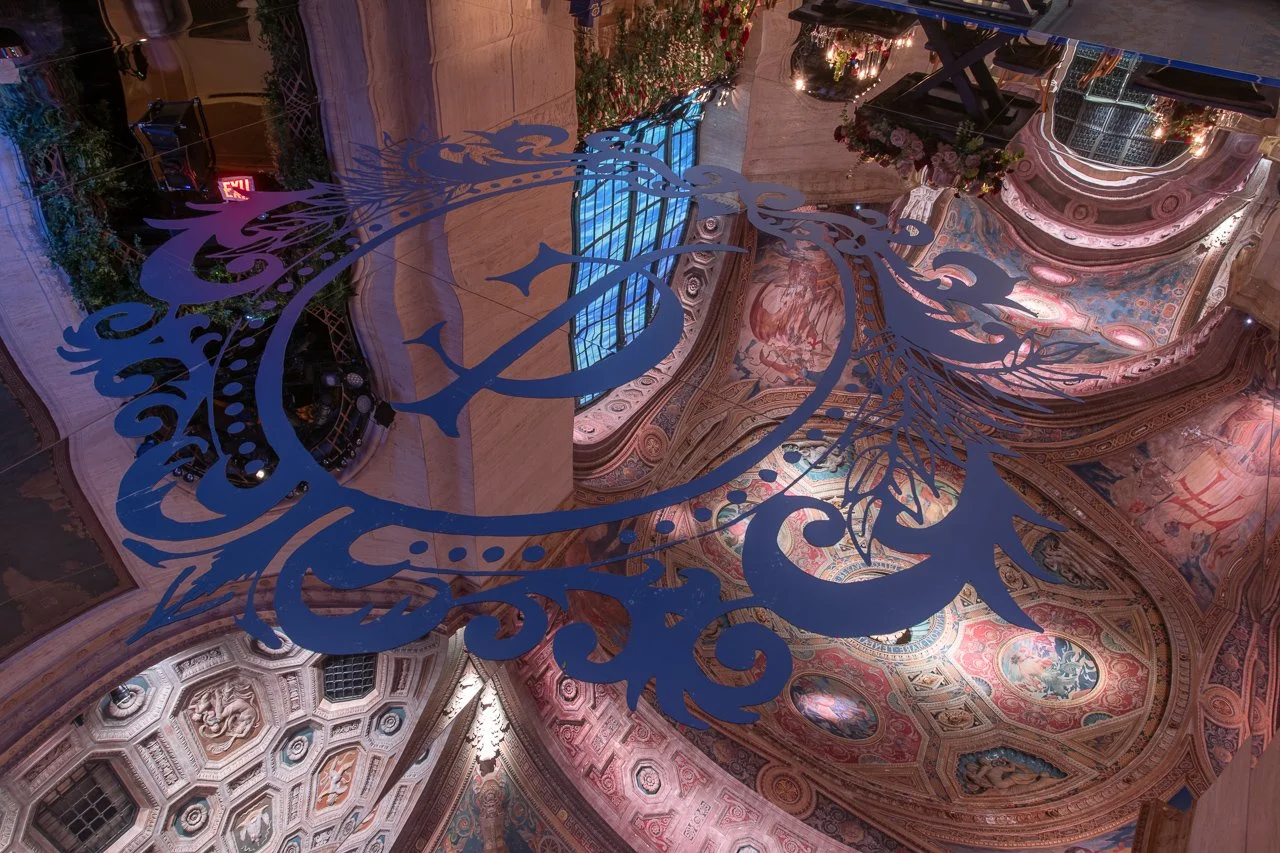 View of an ornate, colorful ceiling inside a historic building, with elaborate mural paintings and intricate architectural details, seen through a large decorative cutout of a winged creature.