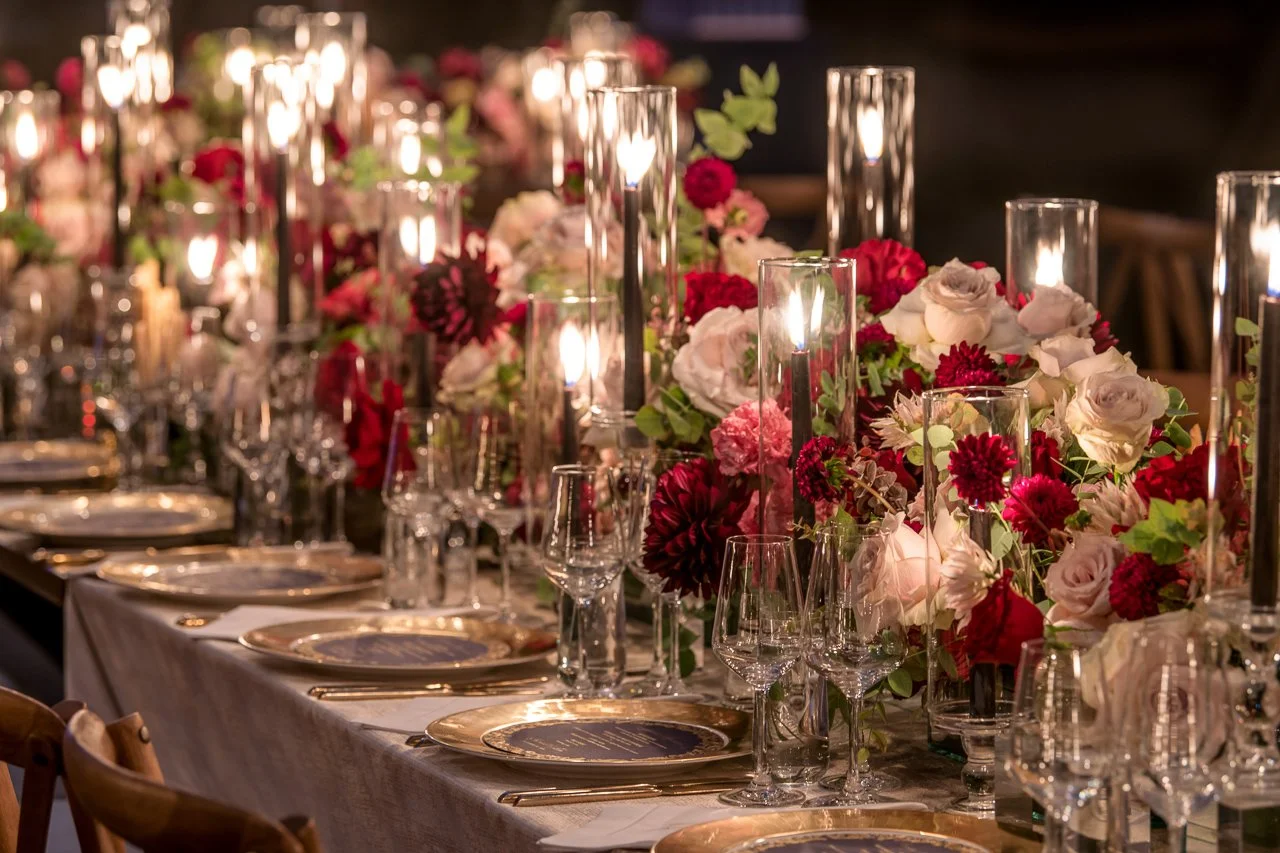 A formal dining table decorated with elegant floral arrangements featuring red, pink, and white flowers, tall glass candle holders with lit candles, and gold-rimmed plates and glassware, set for a special event or celebration.