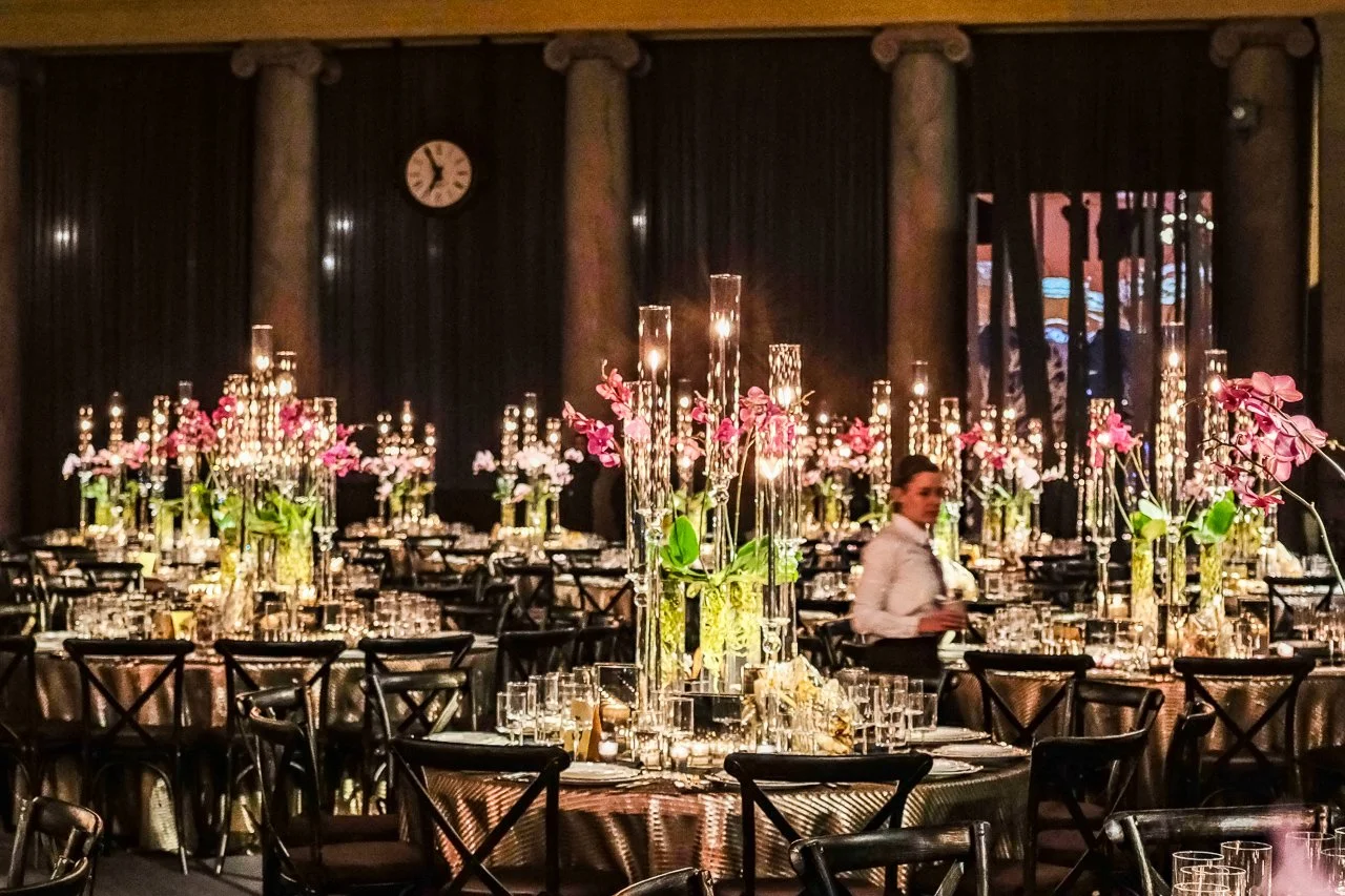 A banquet hall decorated with pink flowers in tall glass vases and lit candles, with black chairs arranged around tables.