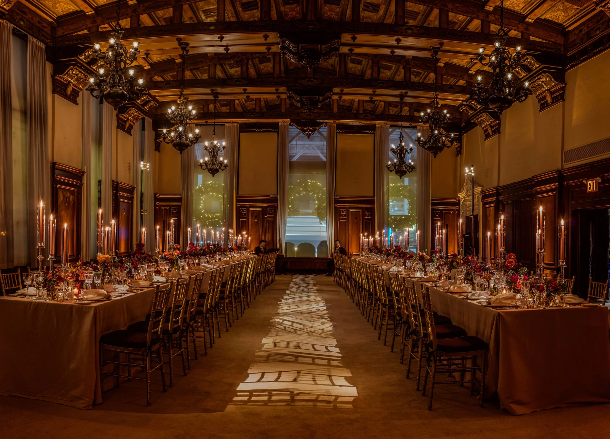 Elegant banquet hall with warm lighting, chandeliers, long tables with gold chairs, floral centerpieces, and candles, set for a formal event.