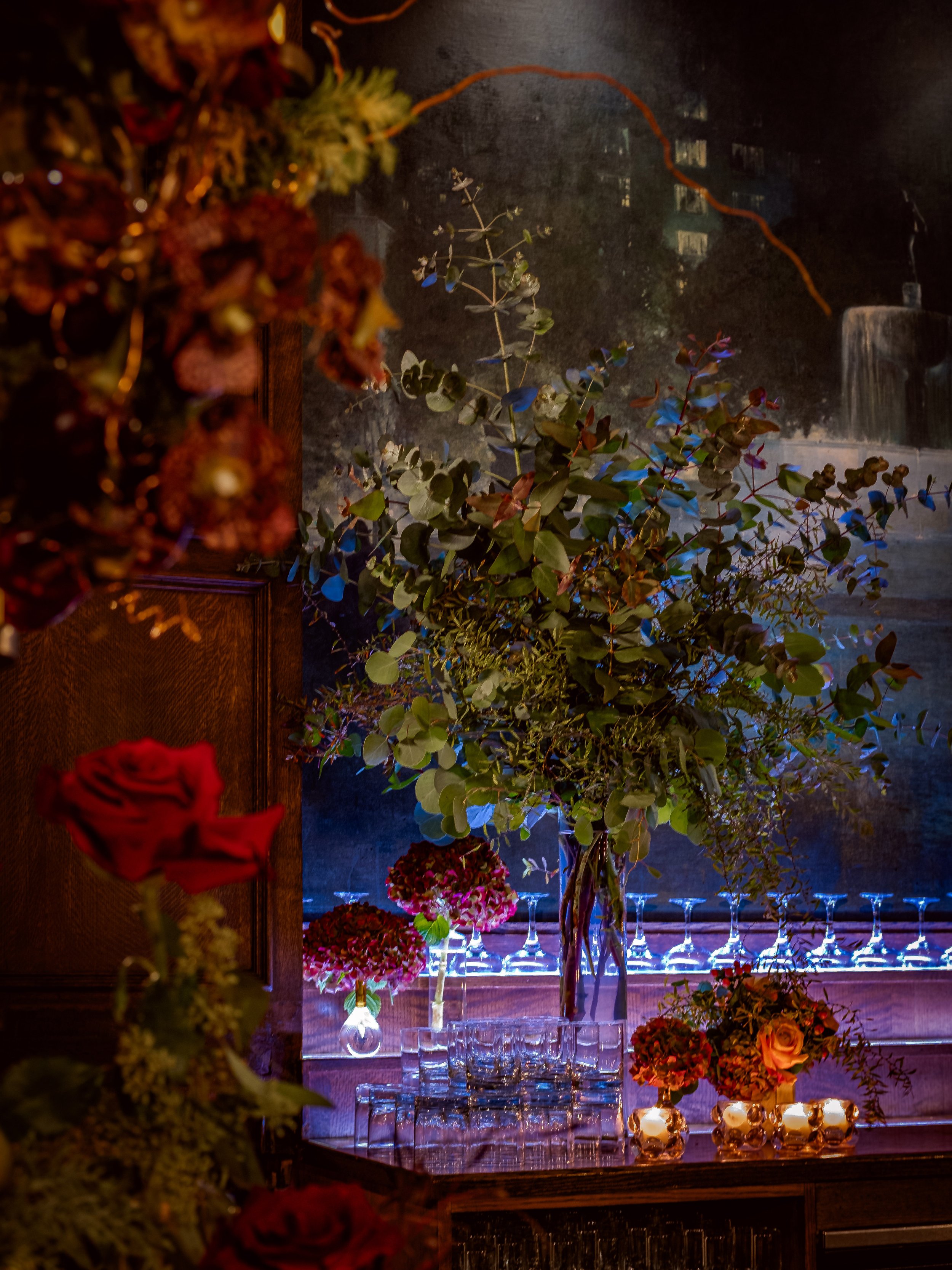 A large glass vase with lush green foliage and branches sits on a bar. Smaller flower arrangements with red, pink, and purple flowers and candles are nearby. Glassware is lined up on the bar, with a dark background and some dim, colorful lighting.