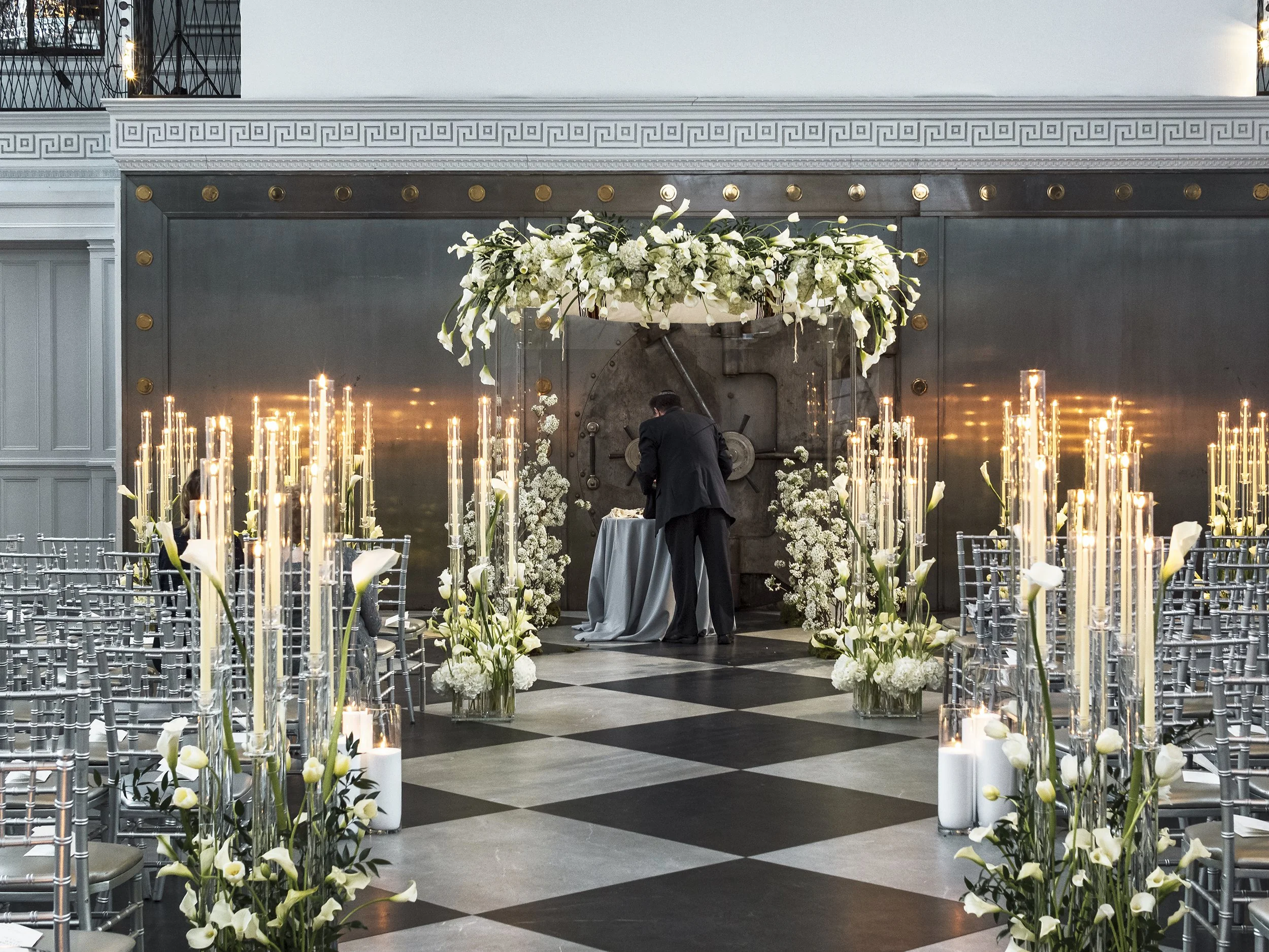 Wedding ceremony setup with silver chairs, white floral arrangements, and candles, featuring a large vault door with a floral arch in the background.
