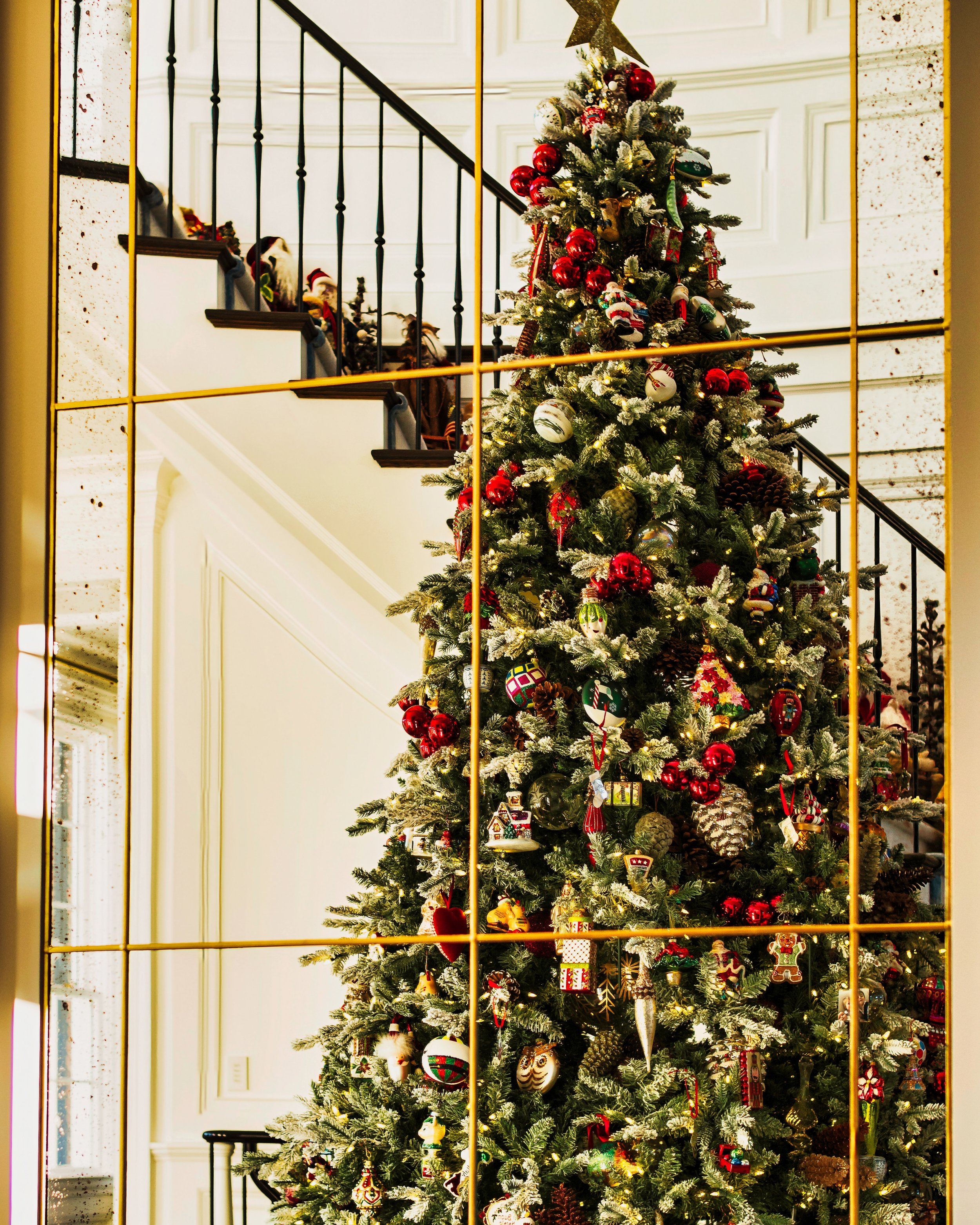 A decorated Christmas tree viewed through a gold lattice mirror frame inside a house with a staircase.