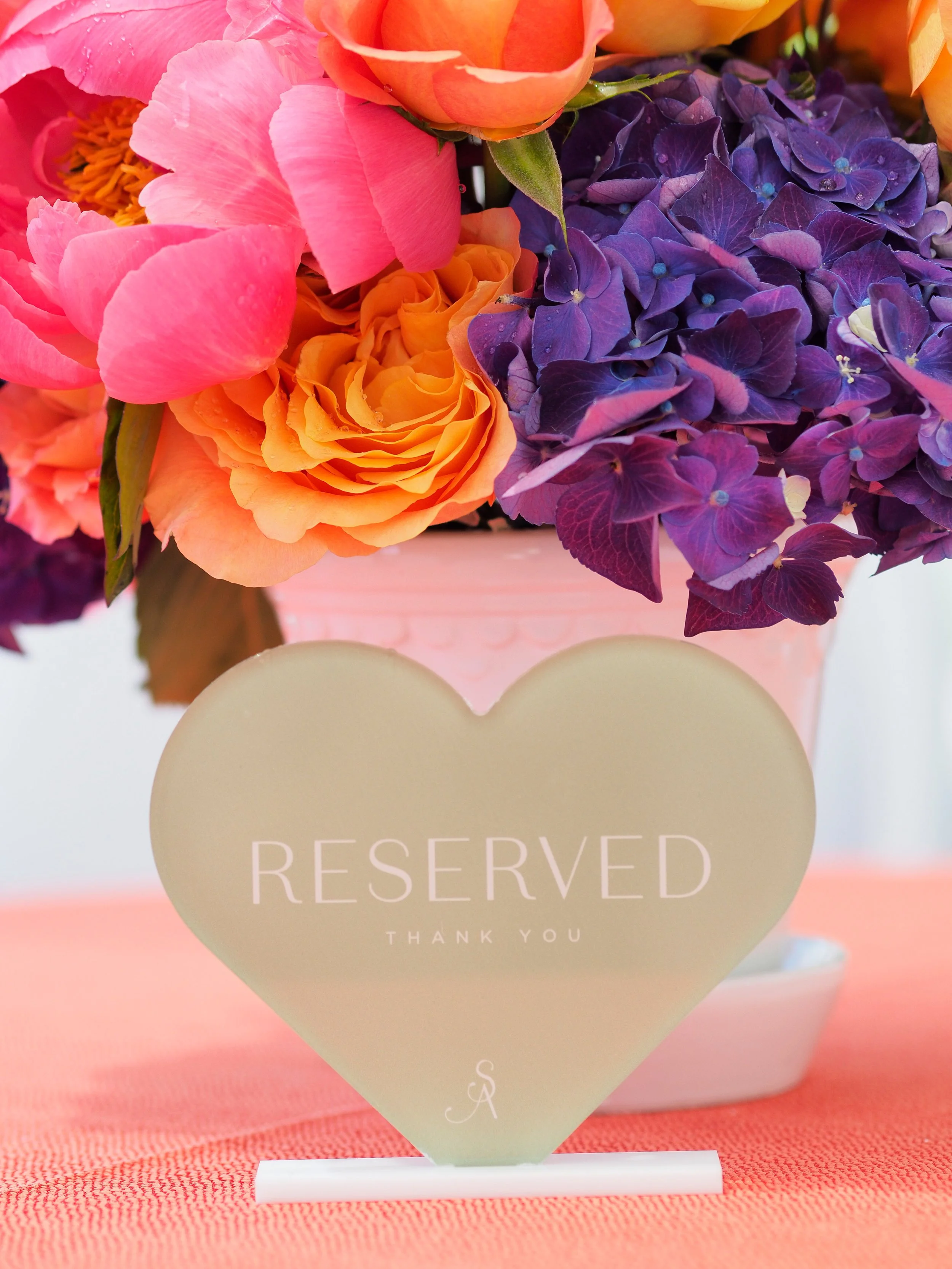 A floral arrangement with pink, orange, and purple flowers behind a heart-shaped 'Reserved' sign on a pink tablecloth.