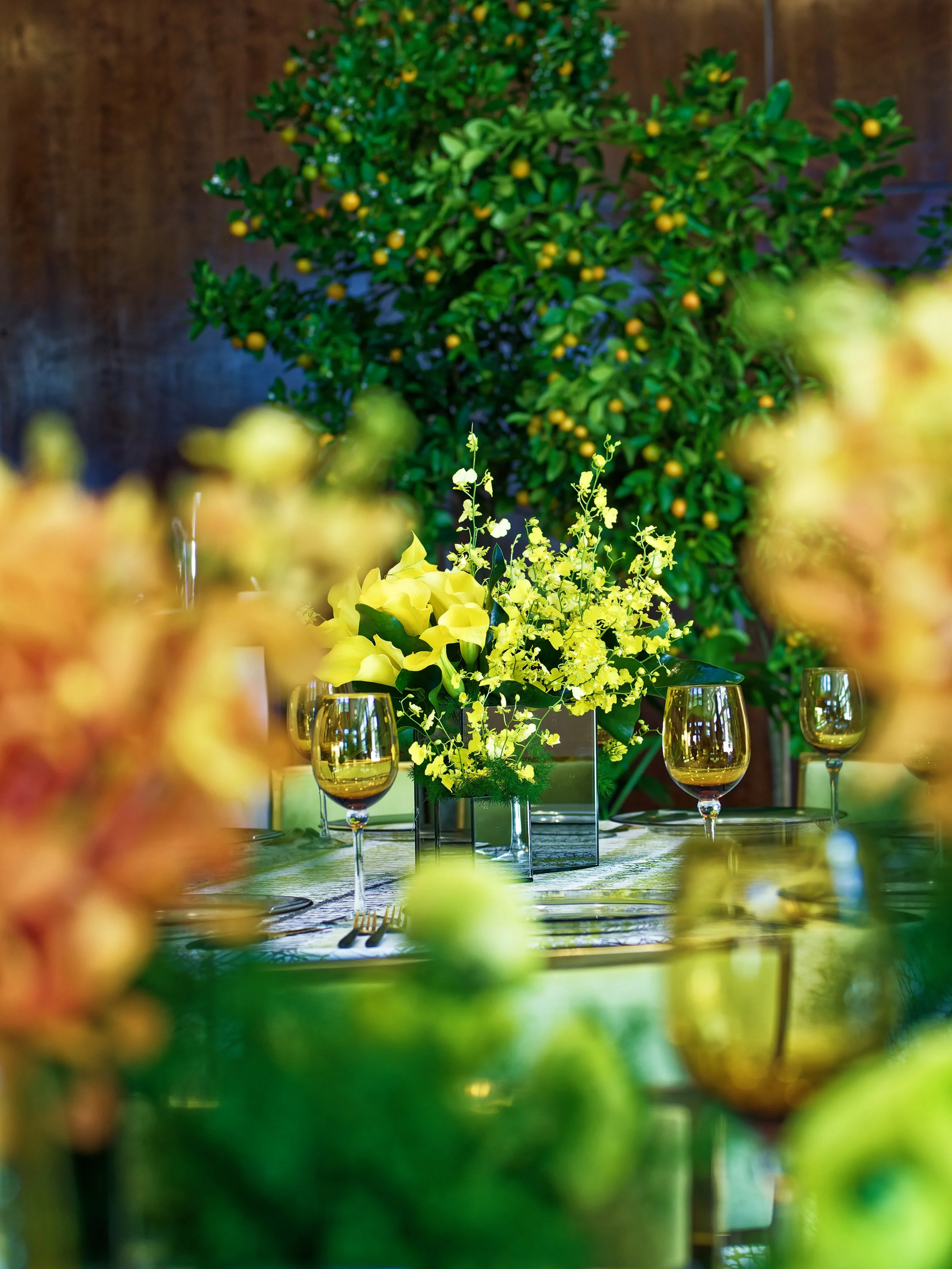 Elegant table setting with yellow floral centerpiece and gold-rimmed glasses, with a lush green plant in the background.