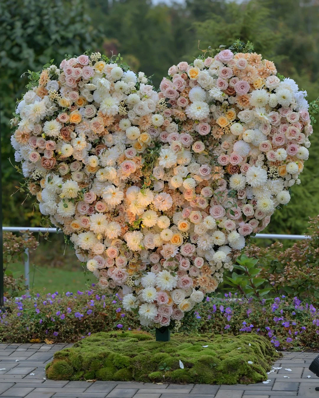 A large heart-shaped floral arrangement made of pink, white, and peach roses and other flowers, placed outdoors on a moss-covered base with purple flowers and greenery in the background.