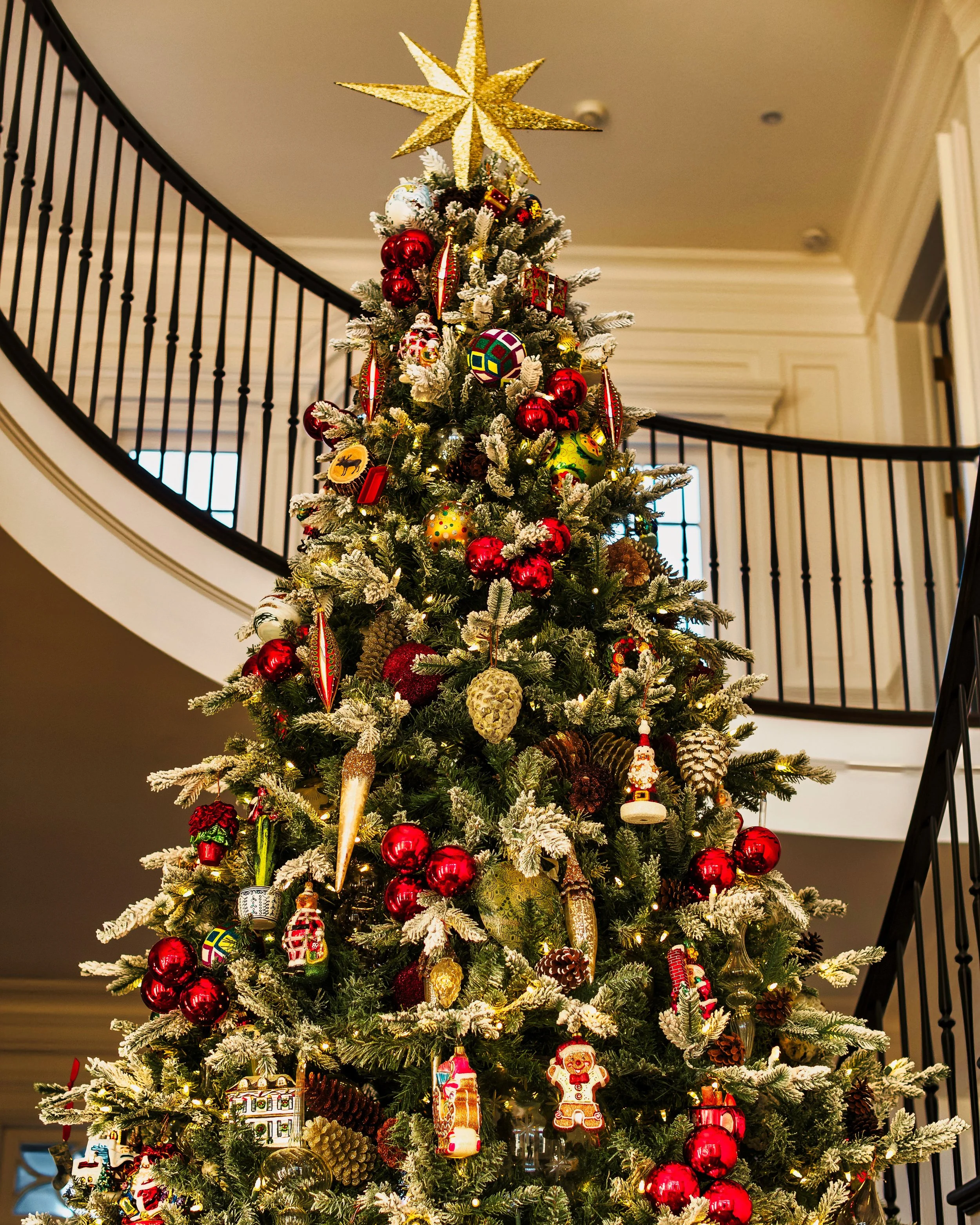 Decorated Christmas tree with red, gold, and colorful ornaments, topped with a large gold star, located in a foyer with a black railing staircase in the background.