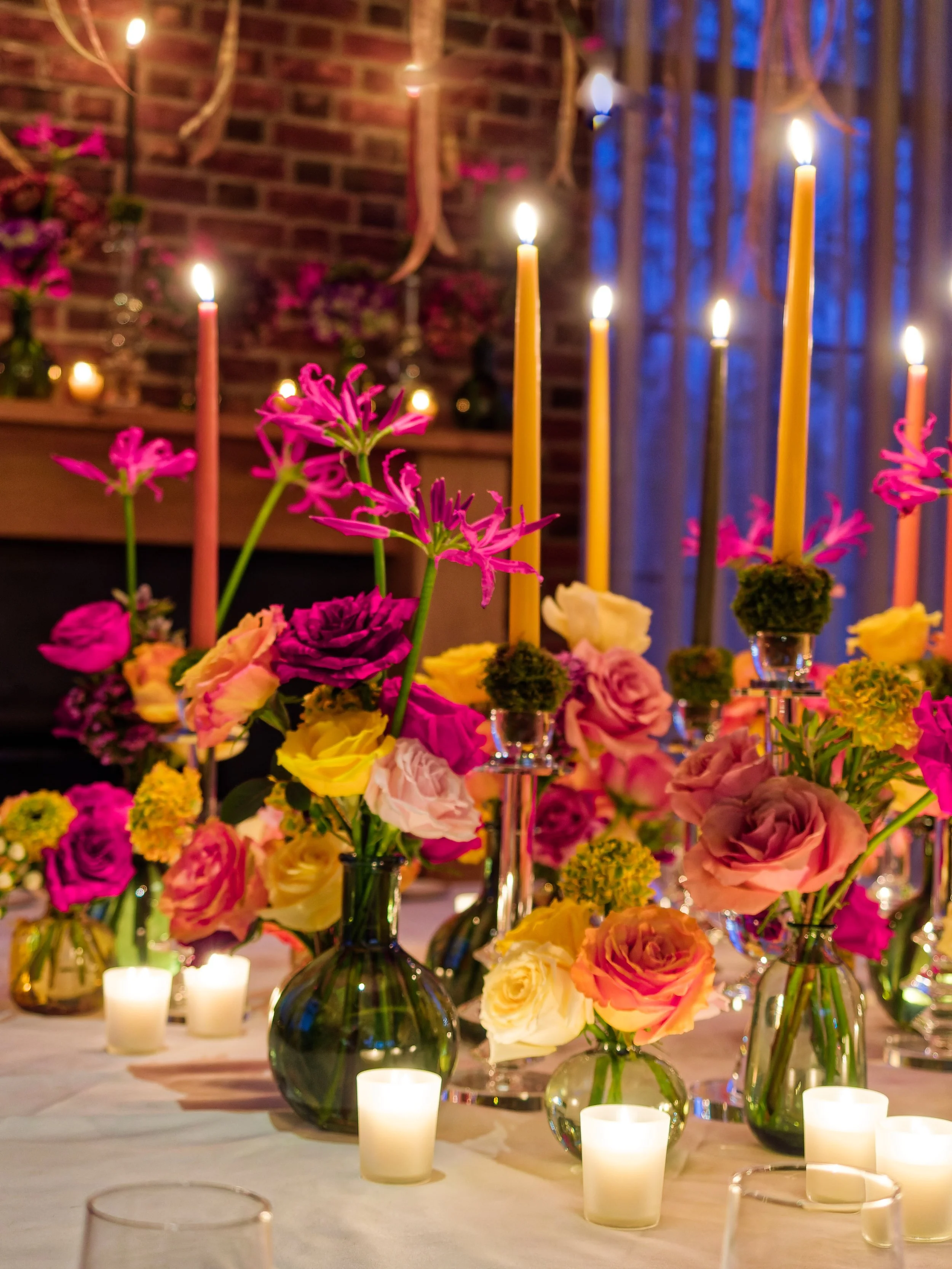 A table decorated with colorful flowers in vases, tall candles, and small candles, with a brick wall and window in the background.
