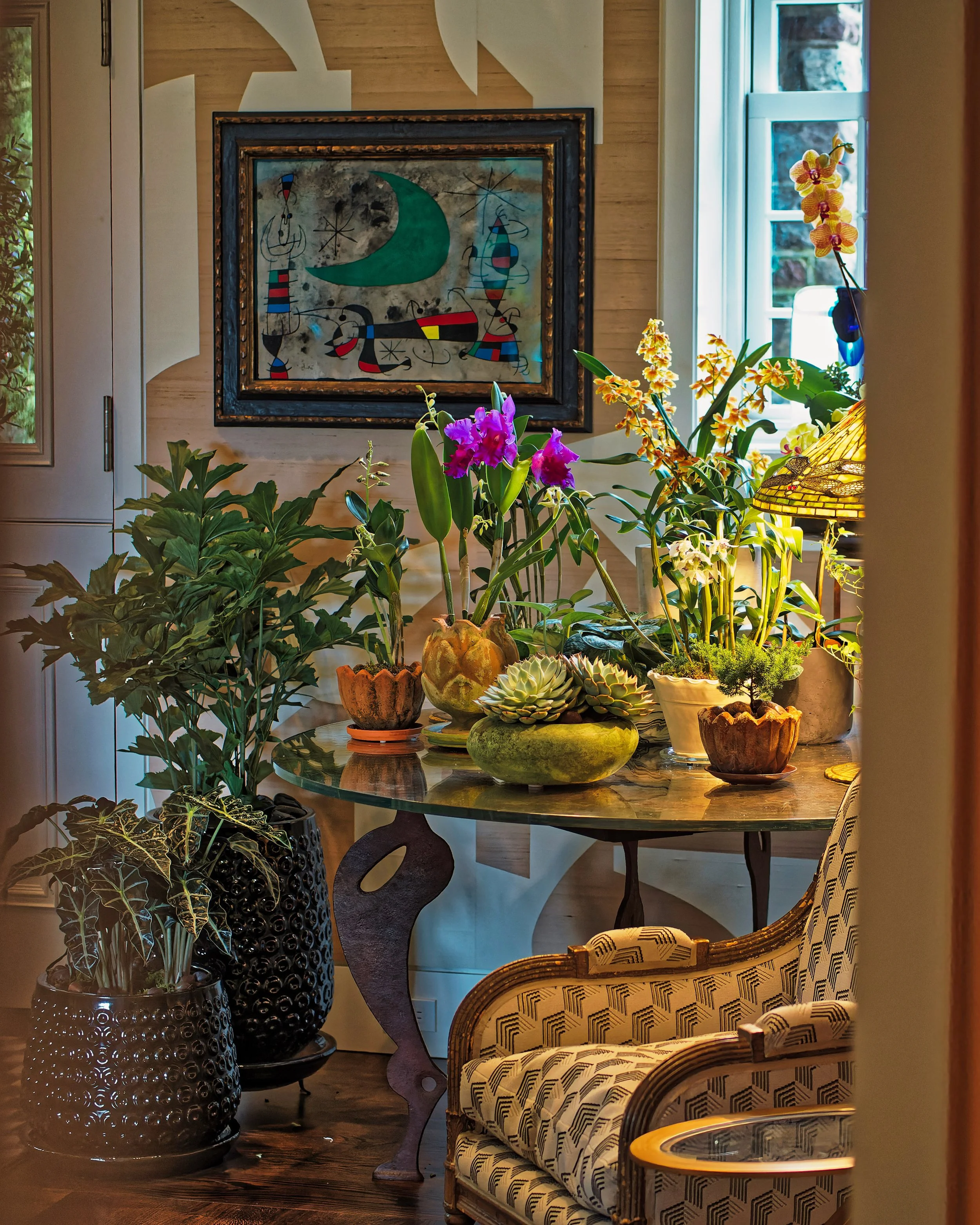 A cozy interior space with a glass-topped table holding potted orchids, succulents, and decorative plants. A patterned armchair is partially visible, and a framed abstract painting hangs on the wall. There is natural light coming through a window.