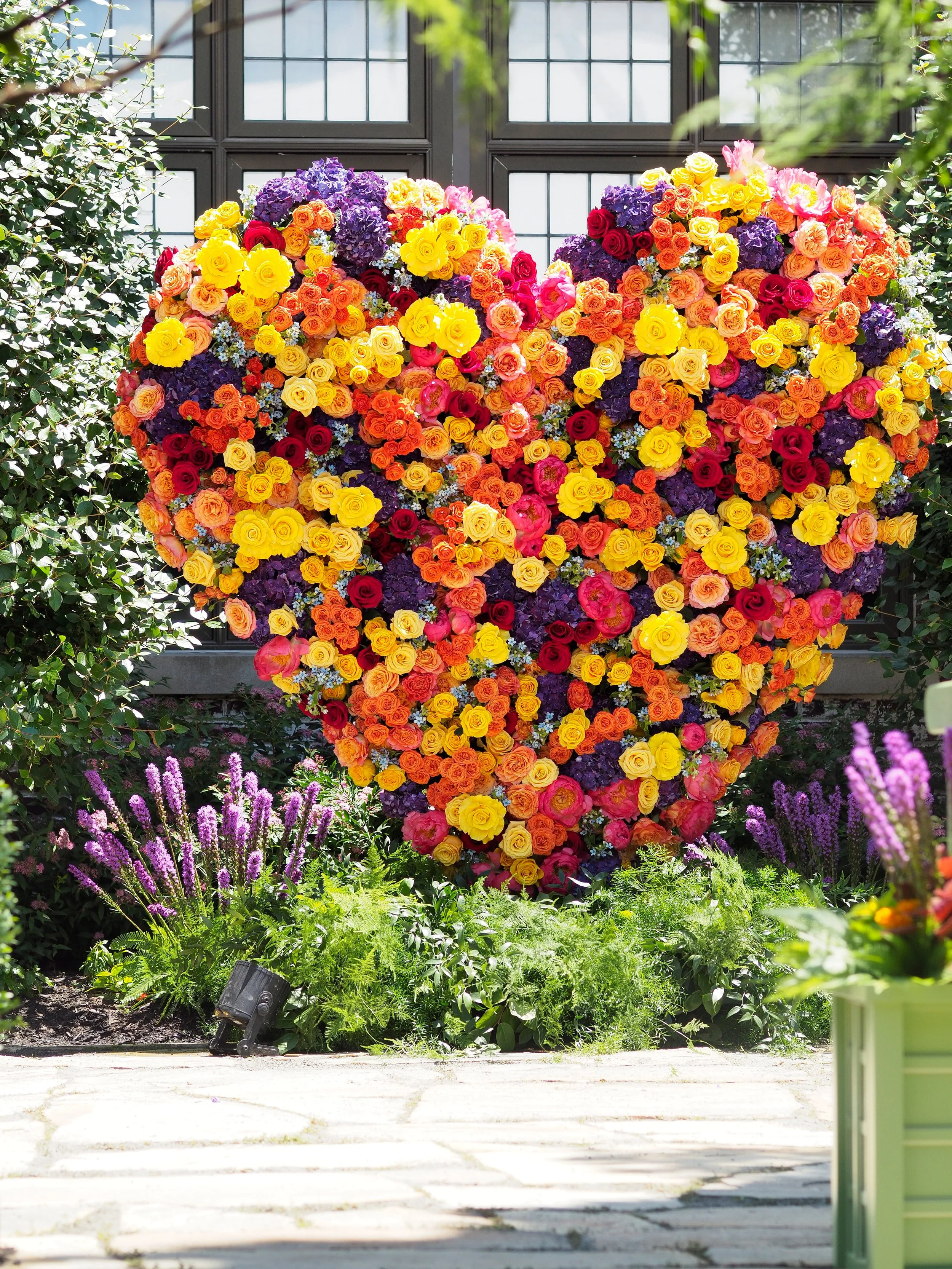 A large heart-shaped floral arrangement made of colorful roses and flowers, displayed outdoors in front of a window with black framing, surrounded by green plants and purple flowers.