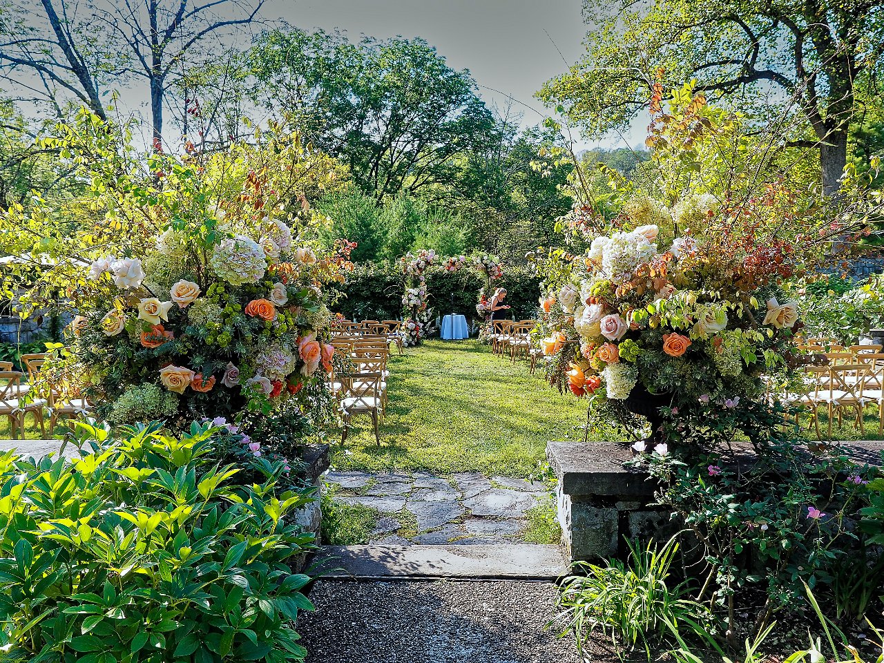 Outdoor wedding ceremony setup with floral arrangements, chairs arranged in rows, and a small table in front of a lush green garden with trees.