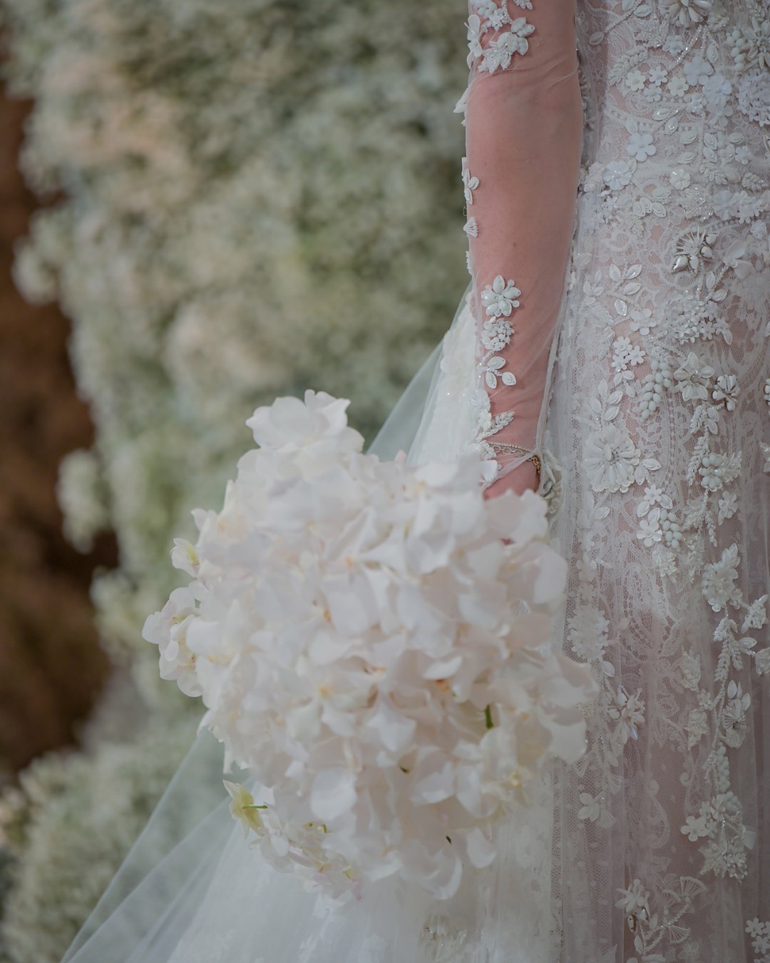 Close-up of a bride holding a bouquet of white flowers, wearing a lace wedding dress with floral appliqué, at an outdoor wedding.
