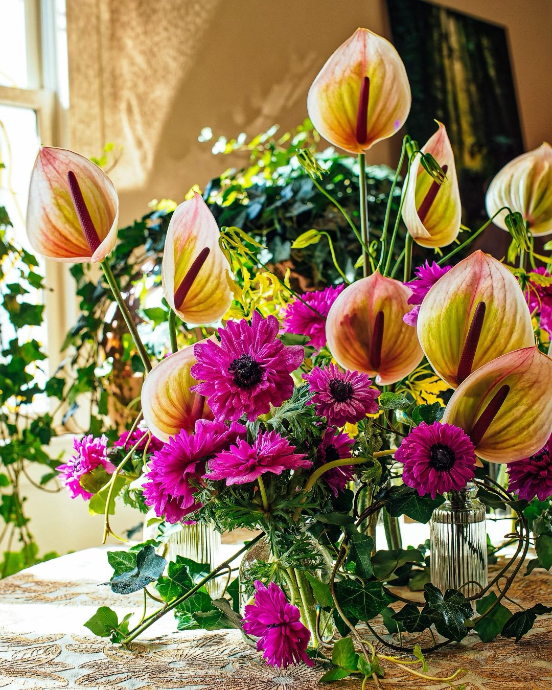 A colorful floral arrangement on a table featuring pink daisies and cream-colored calla lilies with pink and yellow streaks, set against a sunlit background with a window, door, and a blurred painting.