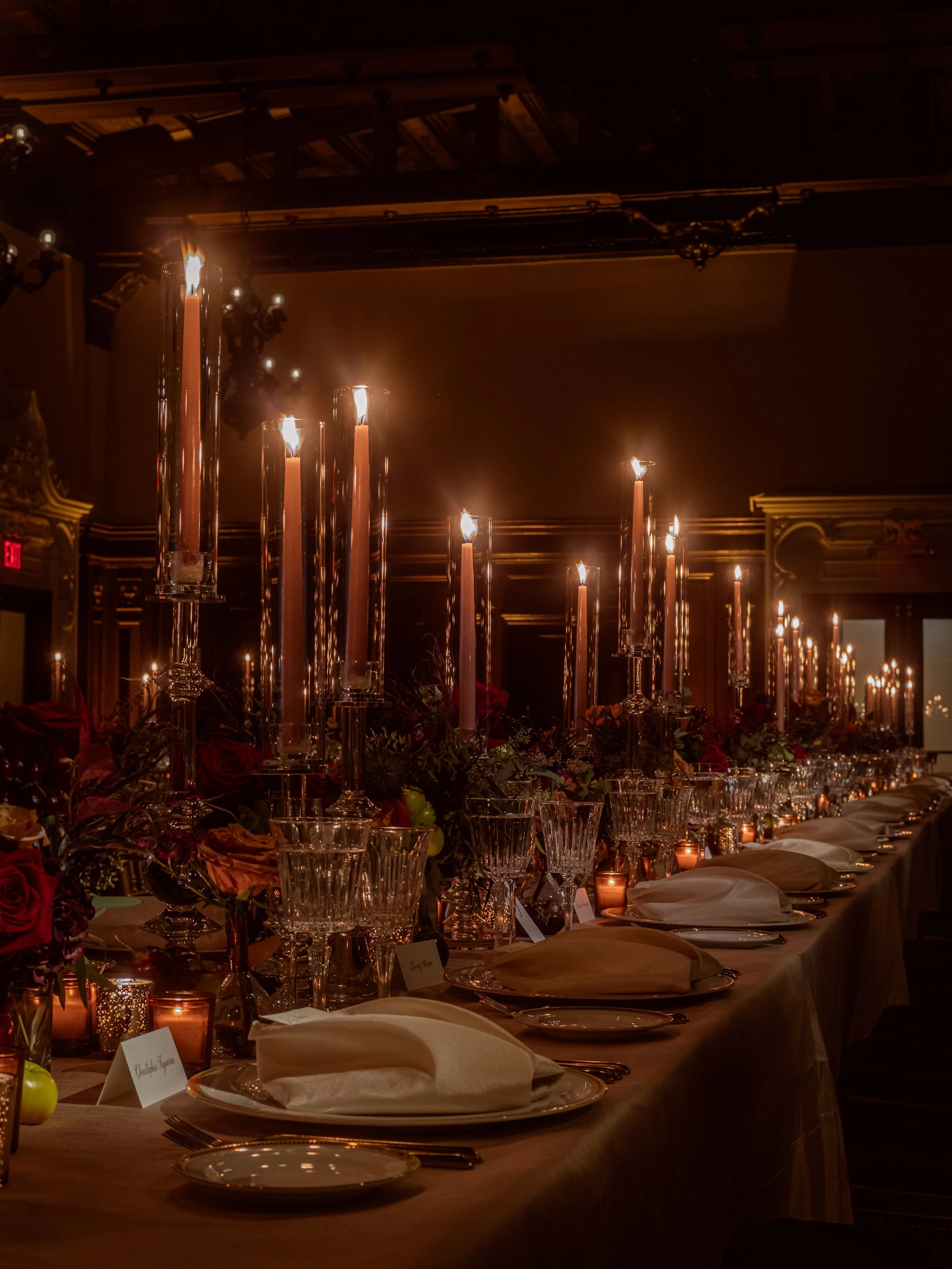 Elegant banquet table decorated with tall candles, floral arrangements, glassware, and neatly folded napkins in a dimly lit room.
