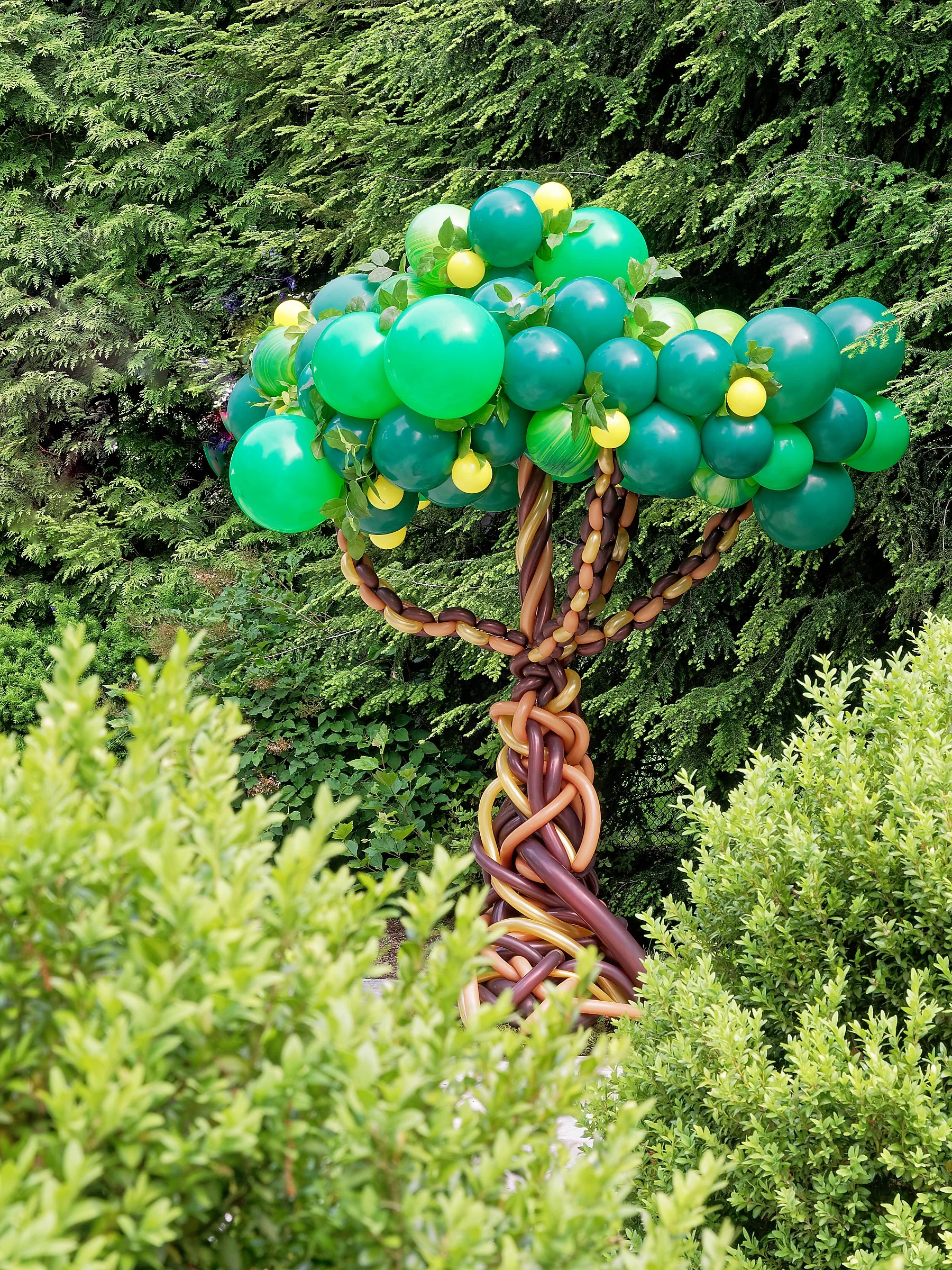 A balloon art sculpture of a tree, with a trunk made of intertwined brown, tan, and purple balloons, and a canopy of green, blue, and yellow balloons, surrounded by green bushes and trees.