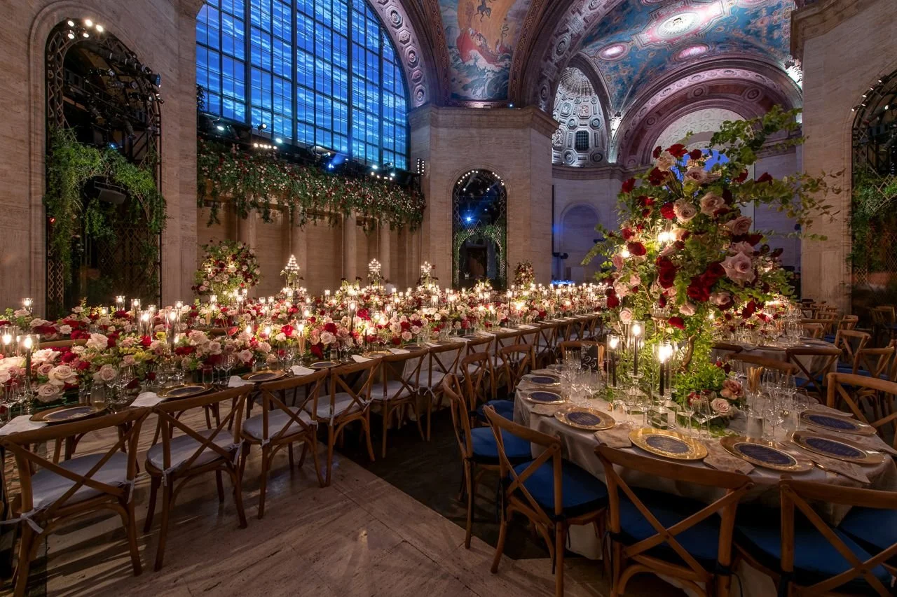 Elegant banquet hall decorated for a wedding or special event with long tables filled with pink and white floral centerpieces, candles, and gold-rimmed plates. The hall features high ceilings with intricate artwork and large stained glass windows.