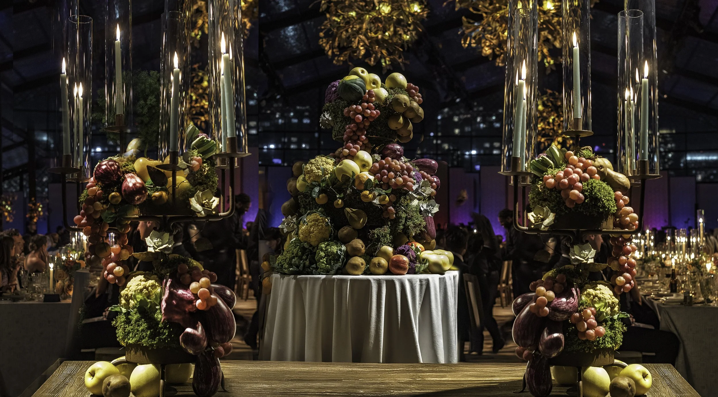 An elegantly decorated event space at night featuring a centerpiece of a fruit and vegetable arrangement on a white tablecloth, surrounded by candelabras with lit candles, with dining tables and guests in the background under a glass roof.