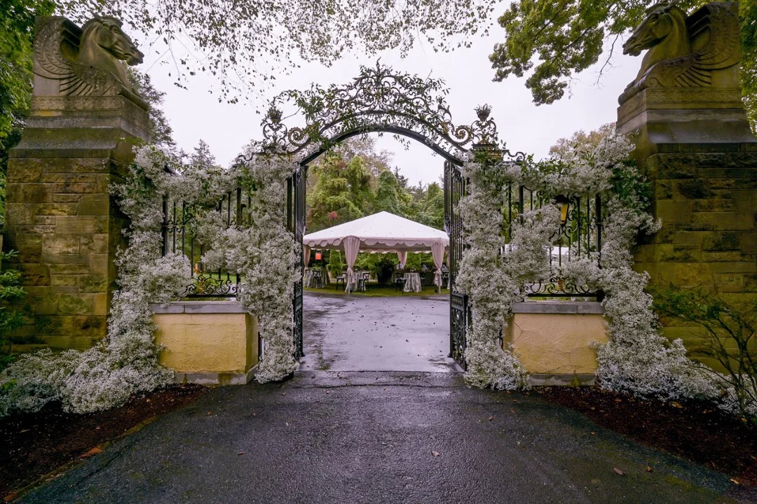 Decorative iron gate decorated with white flowers, leading to a garden with a white canopy tent and tables underneath, surrounded by trees.