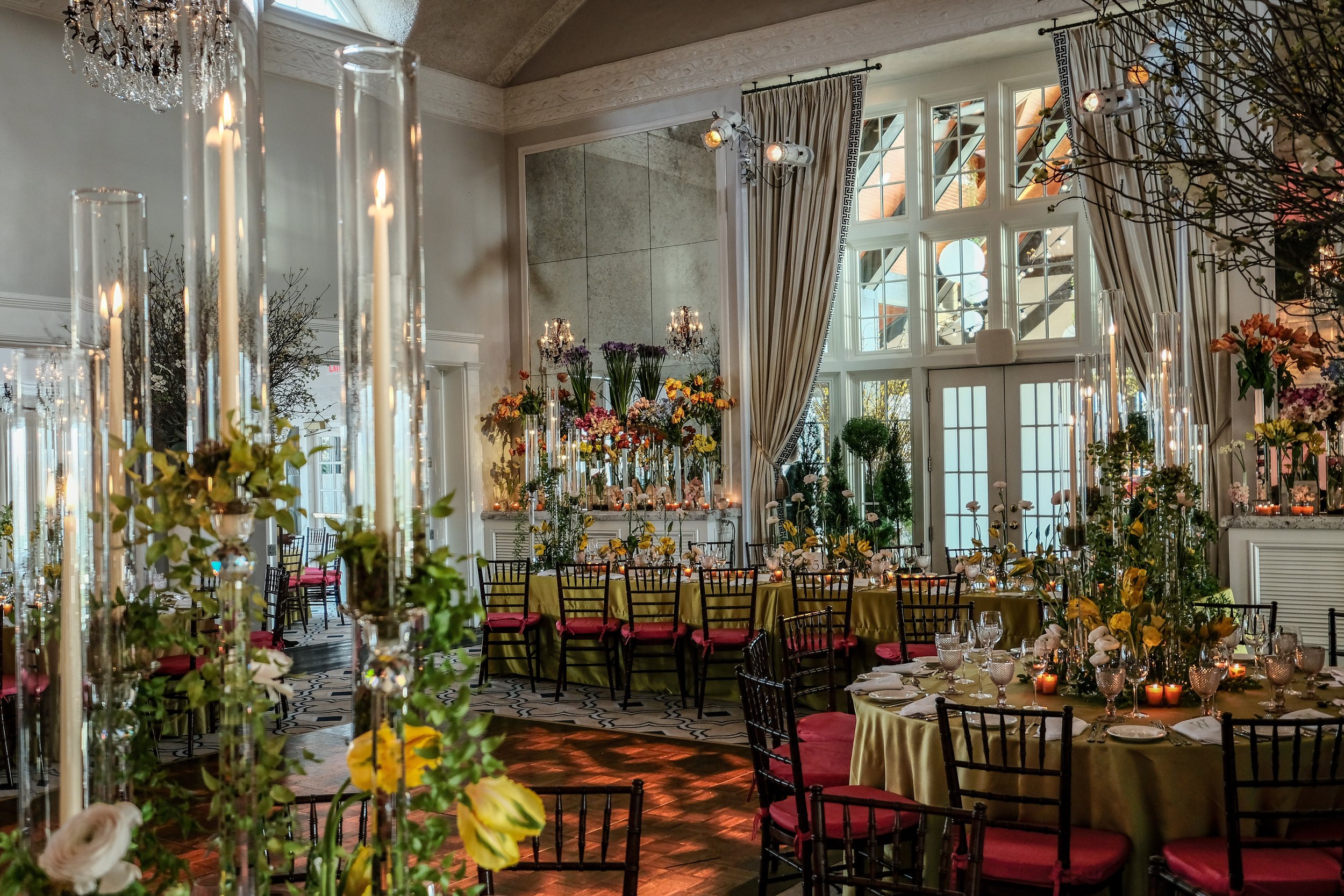 Elegant banquet hall decorated with tall floral centerpieces, candles, and draped curtains, set up for a formal event with round tables, black chairs with pink cushions, and large windows letting in natural light.