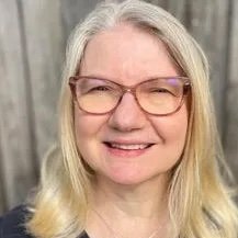 Person with long blond hair wearing glasses and smiling in outdoor setting