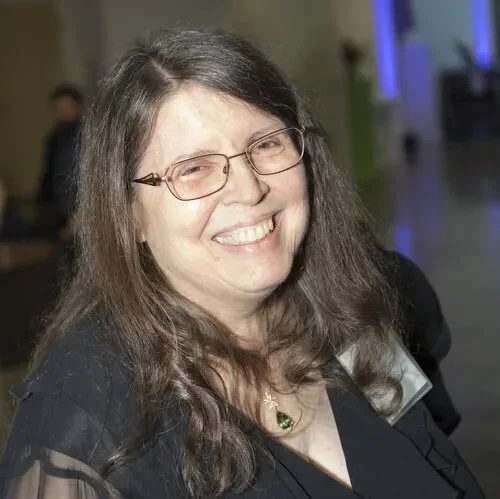Smiling person with glasses and long hair in a dimly lit indoor setting.