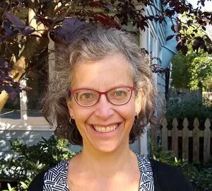 A woman with curly gray hair and red glasses, smiling in an outdoor setting with a picket fence and plants in the background.