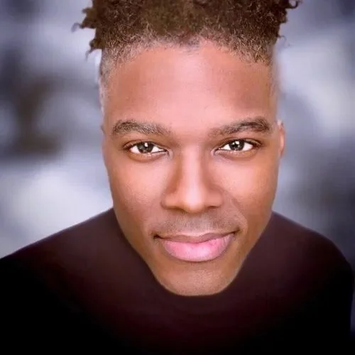 Close-up of a person with short curly hair, wearing a dark shirt, against a blurred background.