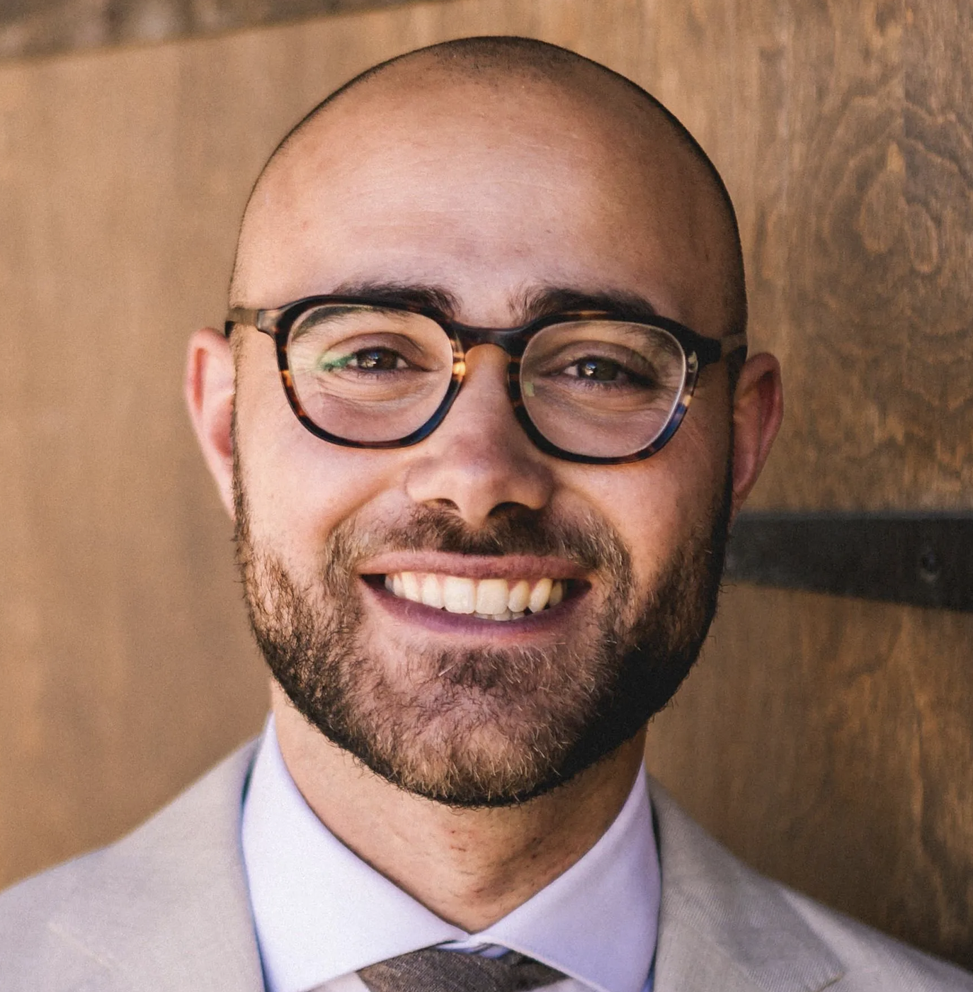 Bald man with glasses smiling, wearing a suit and tie, standing in front of a wooden background.