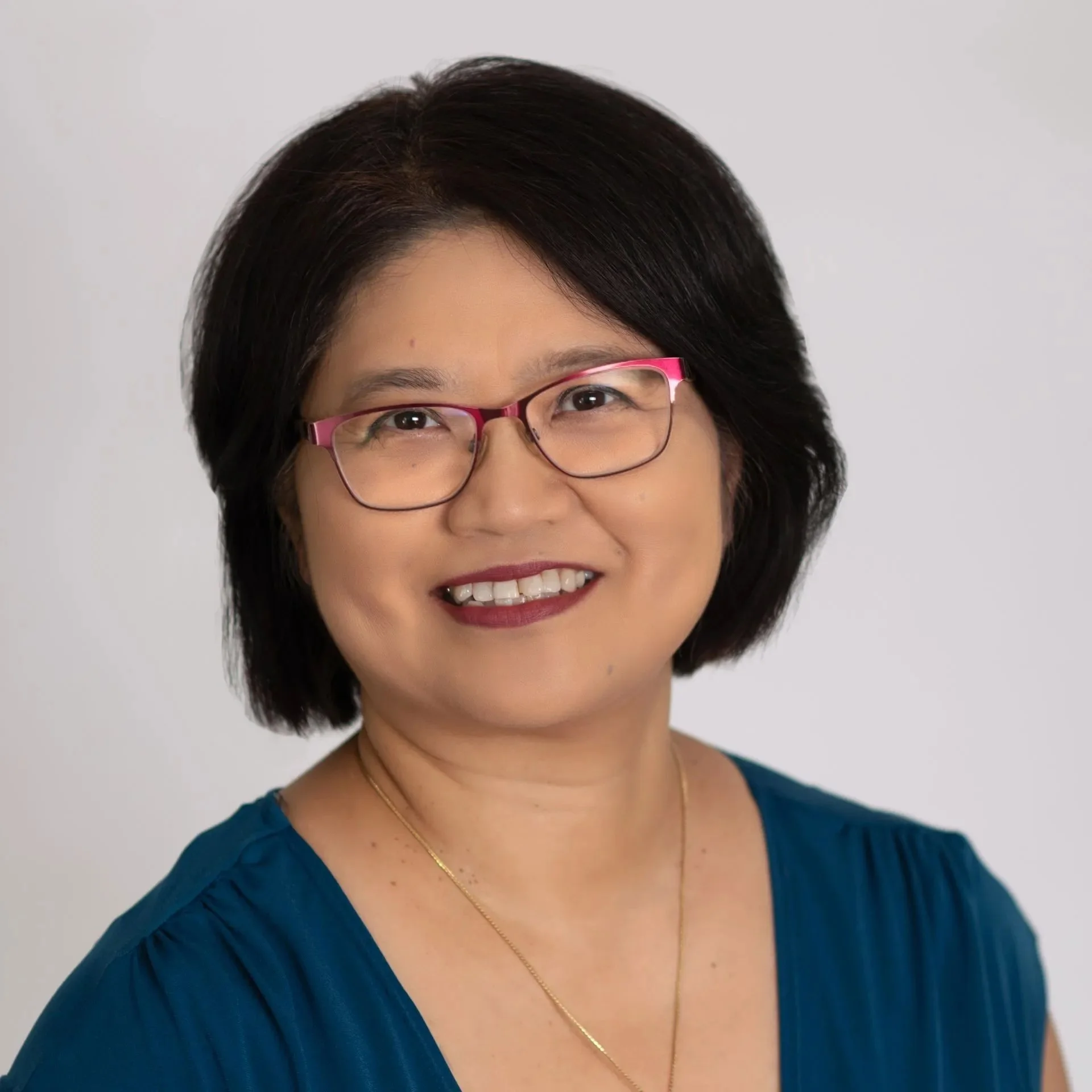 Person with glasses smiling, wearing a blue top and gold necklace, against a light background.