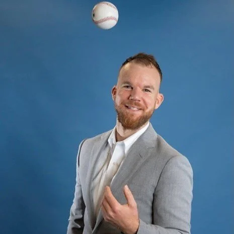 A man in a gray suit smiling and tossing a baseball against a blue background.