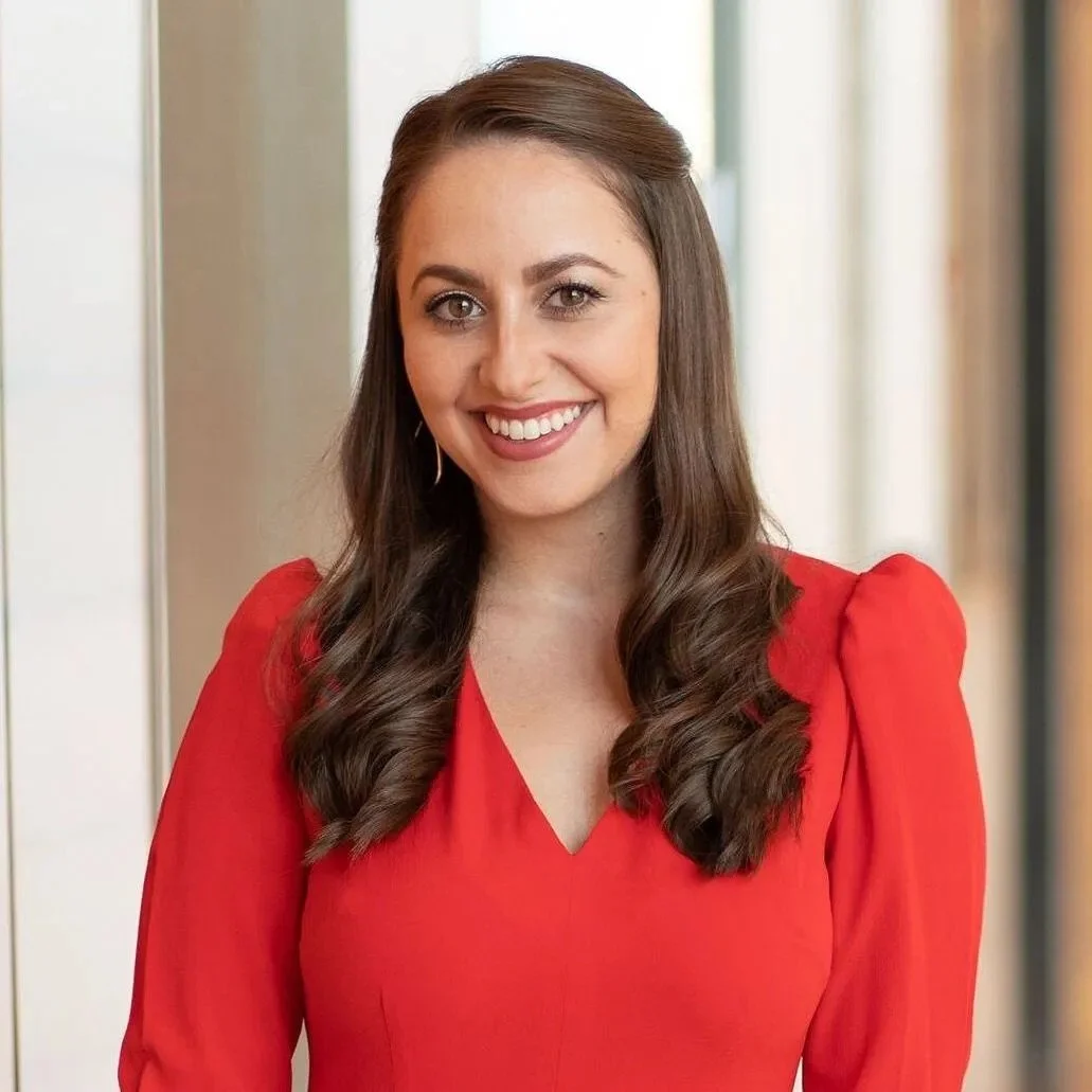 Woman with long brown hair wearing a red dress, smiling in a well-lit indoor setting.