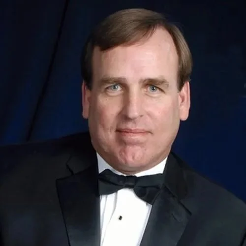 Man in formal attire wearing a black tuxedo with a bow tie, posed against a dark background.