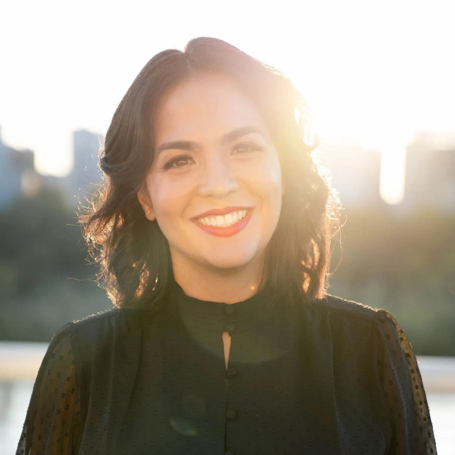 A woman smiling outdoors with the sun shining behind her.