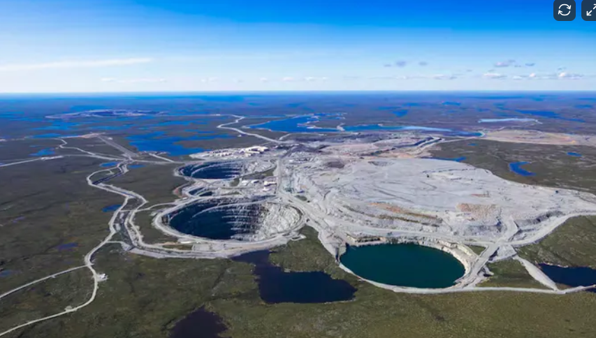 Aerial view of open-pit mining operation with large excavated areas, machinery, and water bodies surrounded by natural landscape and flat terrain.