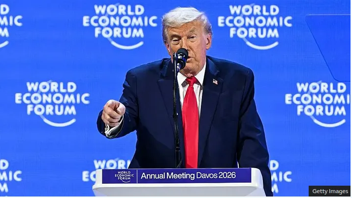 Donald Trump speaking at the World Economic Forum Annual Meeting Davos 2026, standing behind a podium with a blue background displaying the event's name.