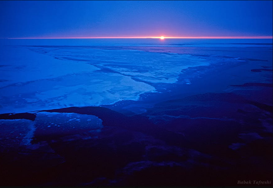 Sunset over icy landscape with frozen water and snow, with a pink and purple horizon and a clear sky.