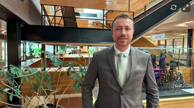 A man in a gray suit and white shirt standing inside a modern building with wooden accents, plants, and a glass staircase.