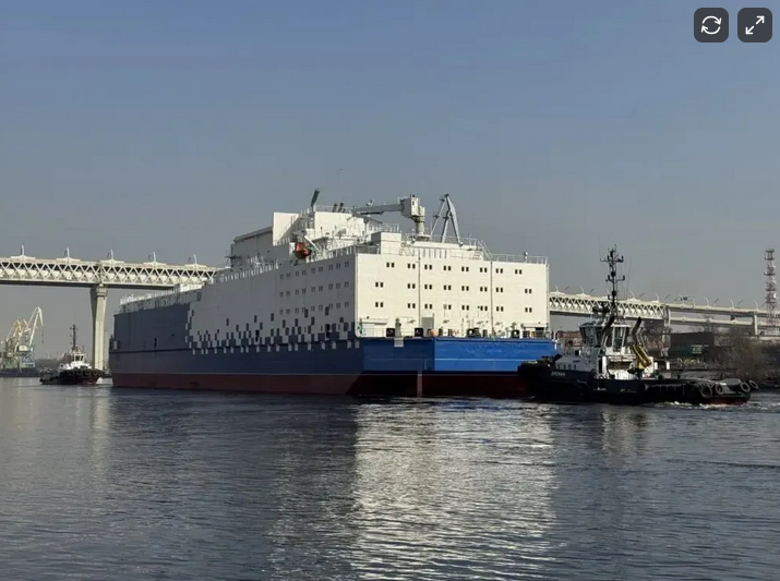 Large white and blue passenger or cargo ship docked in a harbor, with a tugboat assisting it. Bridge structure visible in the background.