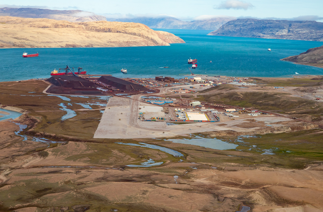 An industrial harbor with ships, storage tanks, and construction areas surrounded by a landscape with rivers, hills, and water in the background.