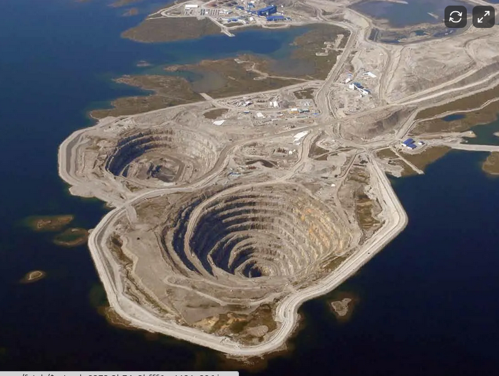 An aerial view of a quarry with large, terraced circular excavations surrounded by roads and infrastructure, located near a body of water.