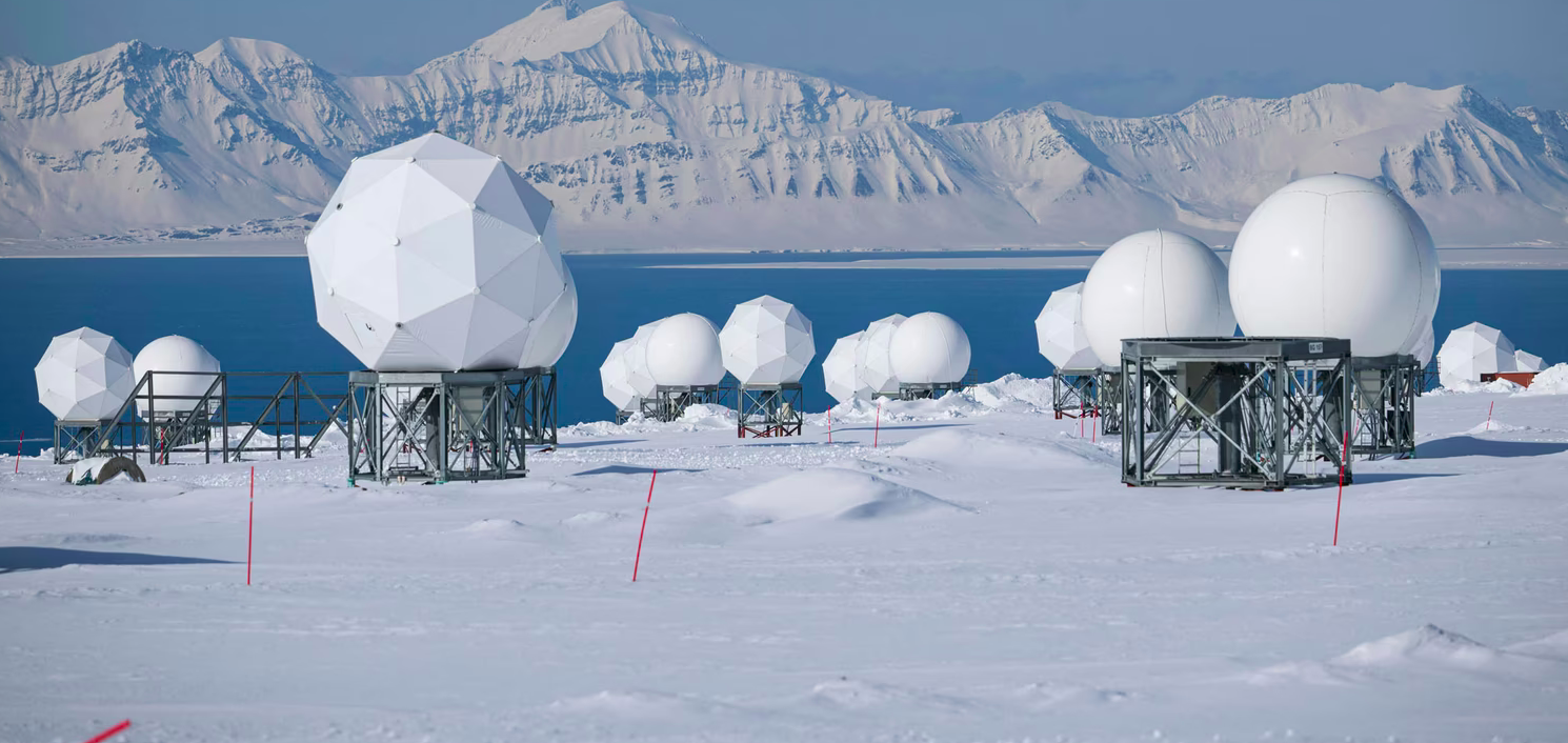 A scientific research station in a polar snowy landscape with radar domes and a mountain range in the background.