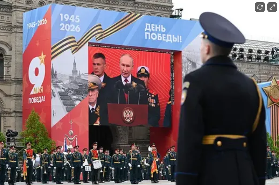 Large outdoor celebration with military personnel and officials on a stage, featuring a large screen displaying Vladimir Putin speaking, with a backdrop commemorating Victory Day on May 9th, marking victory in World War II, with banners showing years 1945-2026 and symbolic decorations.