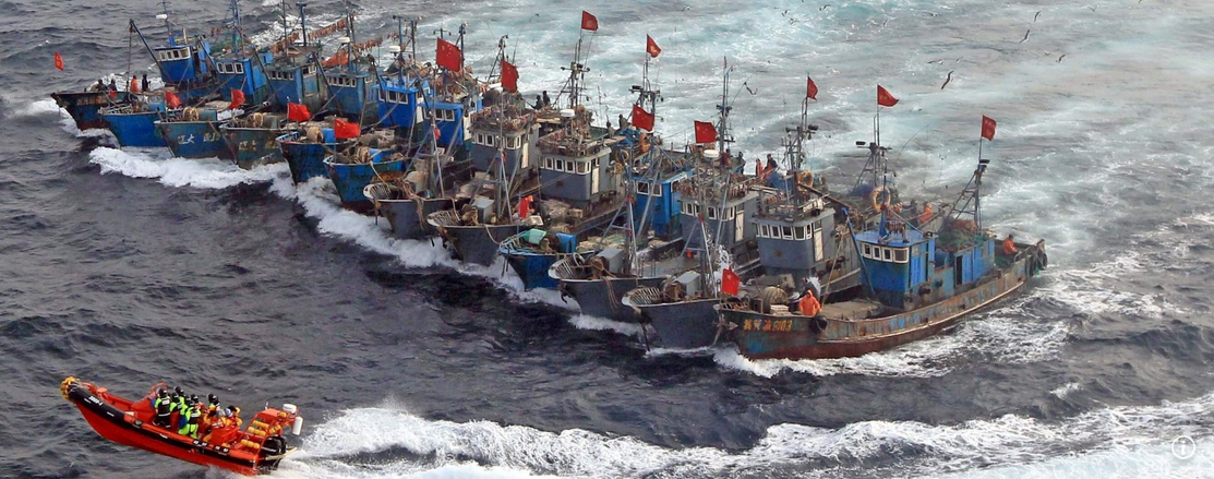A fleet of blue fishing boats with red flags, sailing on choppy waters, accompanied by a small orange rescue boat with personnel on board.
