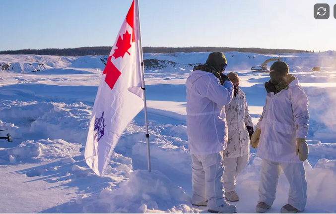 Four people dressed in white winter clothing standing outdoors in a snowy landscape, with a Canadian flag planted in the snow nearby.