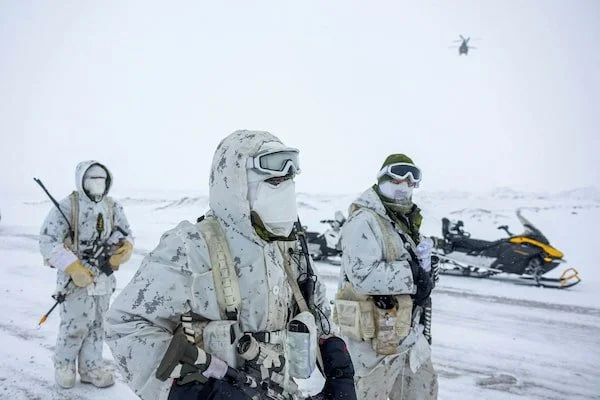 Military personnel in camouflage gear and snow uniforms standing in a snowy landscape, with snowmobiles and a helicopter in the background.