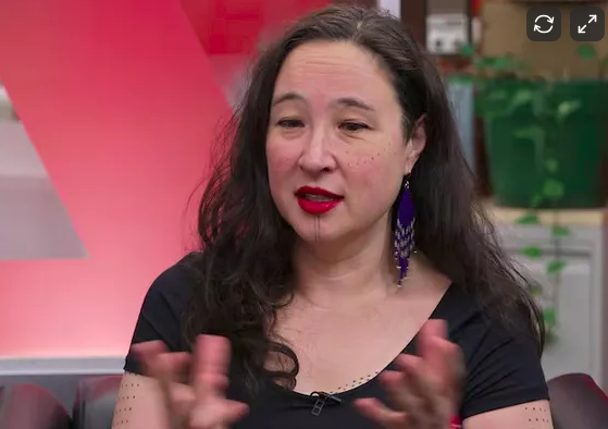 Woman with long dark hair, wearing a black top, red lipstick, and purple earrings, speaking or explaining with hands raised in a studio setting.