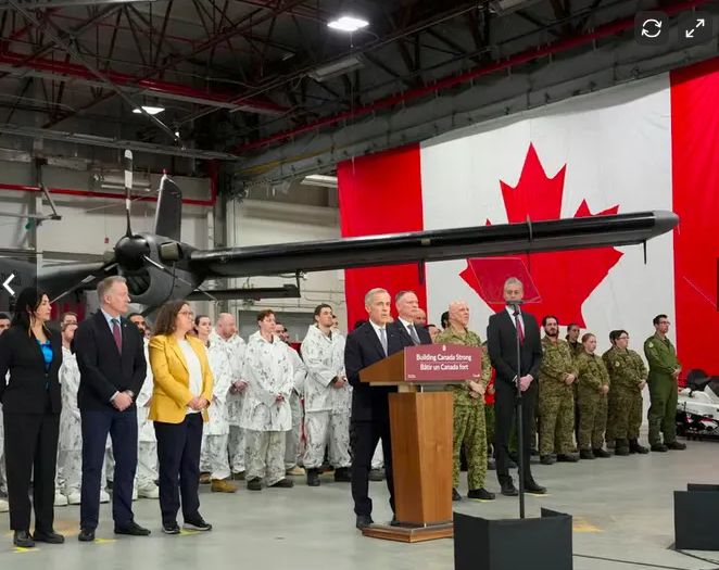 A group of officials and military personnel standing in a hangar with a large Canadian flag hanging on the wall behind them. An aircraft is suspended overhead, and a podium with a speaker is positioned in front.